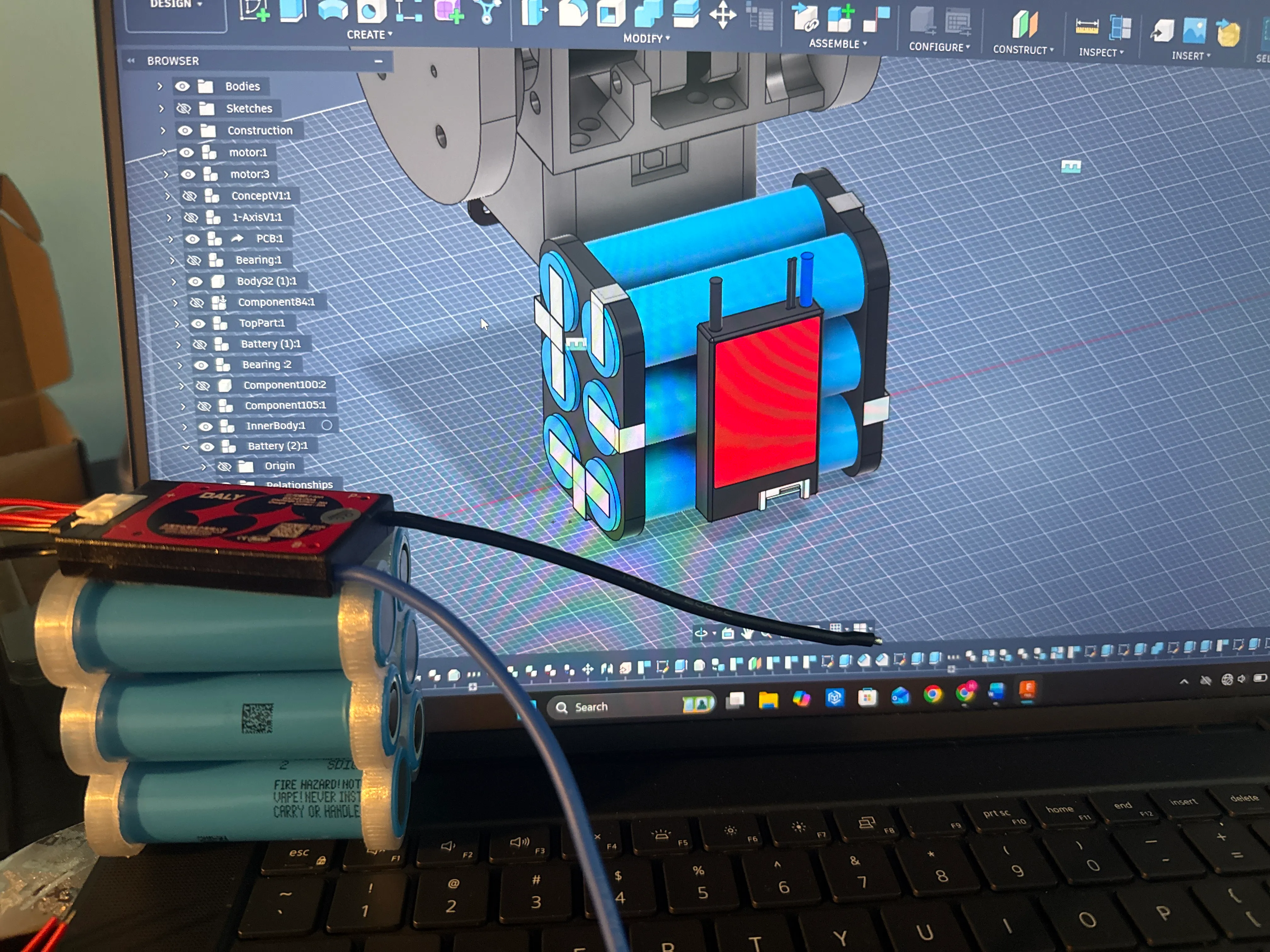 The custom battery pack — 21700 lithium-ion cells in a 3D-printed holder with a red Daly BMS — held in front of a laptop showing the mount's Fusion 360 assembly.