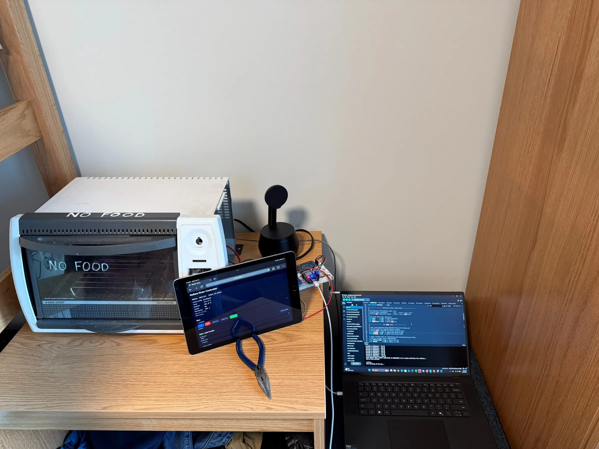 The oven on a desk beside a tablet showing the reflow controller web interface mid-run, and a laptop displaying the firmware source.