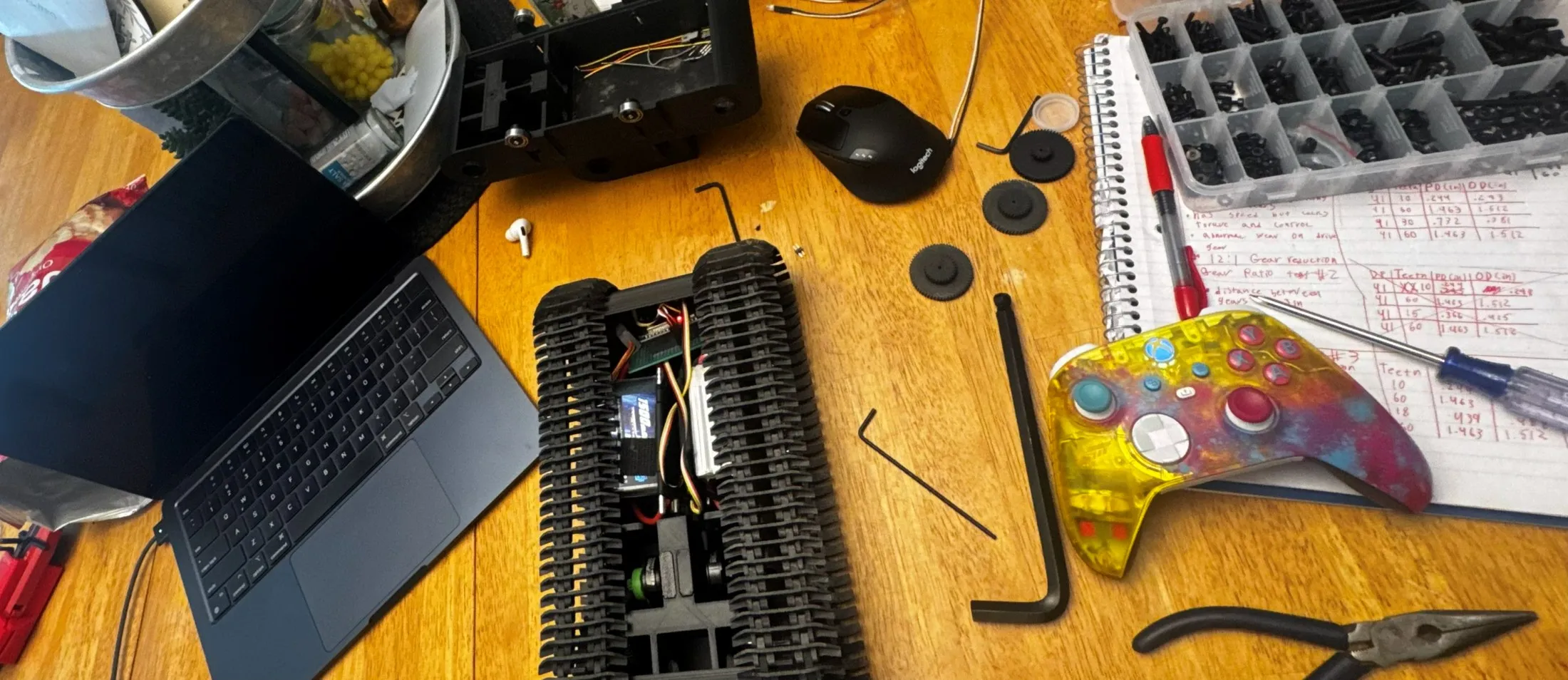 Workbench mid-build — the tracked chassis, a laptop, the printed herringbone gears, a notebook of gear-ratio calculations, and a custom translucent Xbox controller.