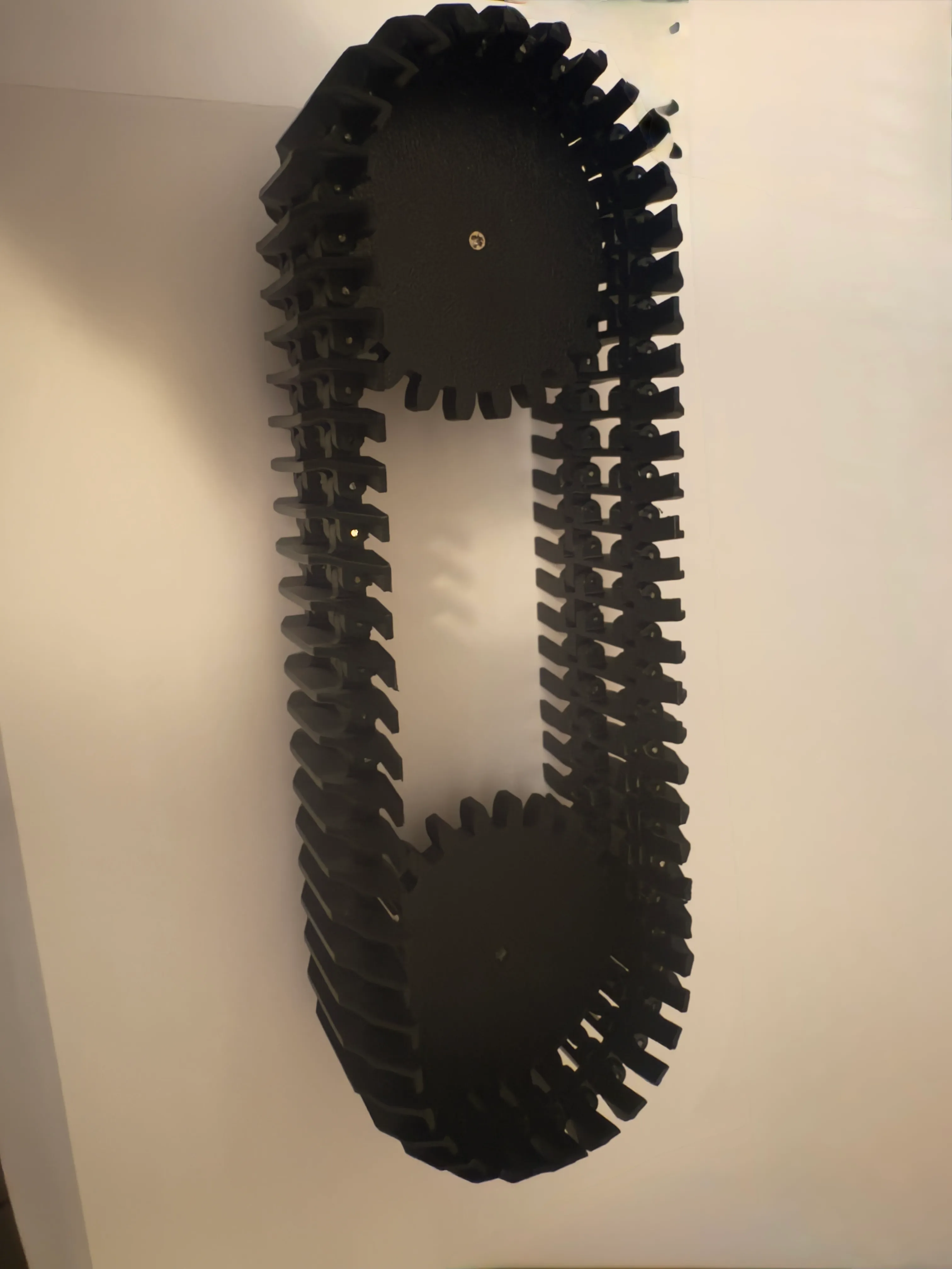 A single 3D-printed track looped over its drive and idler sprockets, held up against a wall — each link hinged on a thin rod.